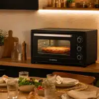 Dyceros Un four de comptoir, dans lequel se trouve un plat au four, est posé sur un plan de travail en bois. Au premier plan, on aperçoit une table à manger dressée avec des assiettes, des verres, des salades et des couverts. Un éclairage chaleureux crée une ambiance conviviale.