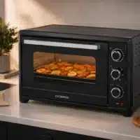 Dyceros Un four de comptoir noir cuit des légumes coupés en rondelles sur une plaque. Le four est équipé de trois boutons de commande et d'une vitre transparente à l'avant. Des ustensiles de cuisine, notamment de l'huile et une plante, sont posés sur le plan de travail à proximité.