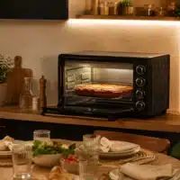 Dyceros Un plan de travail de cuisine sur lequel se trouve un four grille-pain noir dans lequel cuit une lasagne. Au premier plan, une table à manger dressée avec des assiettes, des verres, des serviettes et une salade. La cuisine est baignée d'une lumière chaleureuse et agrémentée de plantes.