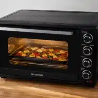 Dyceros Un four de comptoir noir, muni d'une porte vitrée, du logo « DYCEROS » et de quatre boutons de commande, est en train de cuire des légumes coupés en tranches sur une plaque. Le four est posé sur un plan de travail en bois, à côté duquel se trouvent des bocaux et une plante.