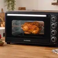 Dyceros Un poulet entier rôtit à la broche dans un four de comptoir noir, au cœur d'une cuisine chaleureuse où des légumes frais et des herbes aromatiques trônent sur le plan de travail en bois.