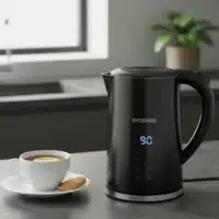 Dyceros Une bouilloire électrique noire avec un écran LED indiquant "90" est posée sur le comptoir d'une cuisine à côté d'une tasse de thé au citron et d'un biscuit sur une soucoupe. Connue pour sa fabrication française, cette bouilloire est à la fois robuste et durable. Une plante en pot et une fenêtre sont visibles derrière.