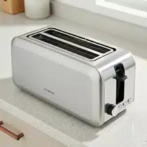 Dyceros Un grille-pain DYERDS argenté à deux fentes, connu pour son design robuste, est posé sur un plan de travail de cuisine de couleur claire près d'une fenêtre. Il comporte un levier et des boutons de commande sur la face avant.