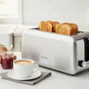 Dyceros Un grille-pain en argent robuste contenant quatre tranches de pain grillé est posé sur le comptoir d'une cuisine à côté d'une tasse de café, d'une assiette, d'un croissant, d'une miche de pain et d'un pot de confiture. Une fenêtre lumineuse se trouve à l'arrière-plan.