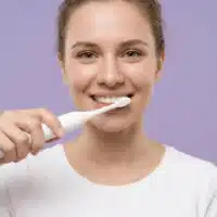 Sensipulse_femme_blanc Dyceros Une jeune femme en t-shirt blanc sourit tout en se brossant les dents avec une brosse à dents électrique durable sur un fond violet clair.