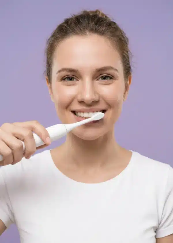 Sensipulse_femme_blanc Dyceros Une jeune femme en t-shirt blanc sourit tout en se brossant les dents avec une brosse à dents électrique durable sur un fond violet clair.