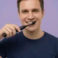 Sensipulse_homme_vieux_noir Dyceros Un homme vêtu d'un t-shirt bleu marine sourit tout en se brossant les dents avec une brosse à dents électrique noire robuste sur un fond violet clair.