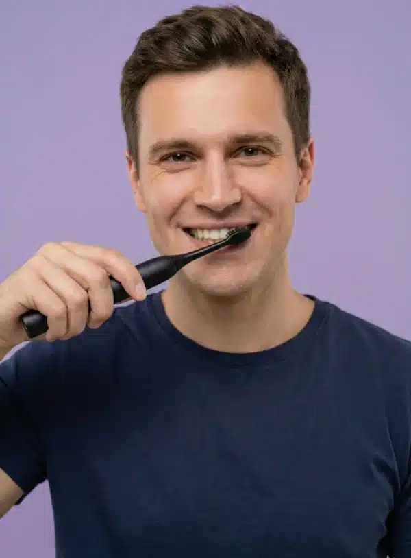 Sensipulse_homme_vieux_noir Dyceros Un homme vêtu d'un t-shirt bleu marine sourit tout en se brossant les dents avec une brosse à dents électrique noire robuste sur un fond violet clair.