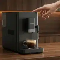Dyceros Une main s'apprête à appuyer sur le bouton d'une machine à espresso moderne DYCEROS noire, qui prépare une tasse de café dans une tasse en verre transparent posée sur son plateau d'égouttage. L'arrière-plan présente des armoires de cuisine en bois.