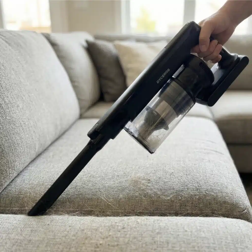 Dyceros Une personne utilise un aspirateur à main noir doté d'un embout étroit pour nettoyer les interstices d'un canapé en tissu gris clair dans un salon lumineux.