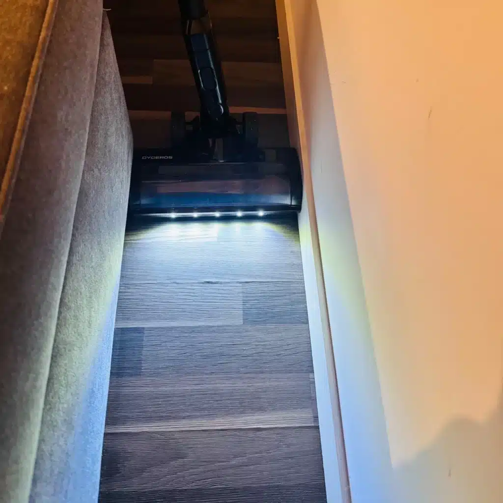 Dyceros Un aspirateur équipé d'une lampe LED intégrée nettoie un espace étroit entre un canapé et un mur sur un sol en bois.