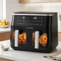 Dyceros Une friteuse Dyceros ClearFry Duo 9L avec deux compartiments de 4,5L et des fenêtres de visualisation est posée sur le comptoir d'une cuisine, faisant cuire des frites d'un côté et du poulet assaisonné de l'autre. L'écran numérique indique les réglages de température.