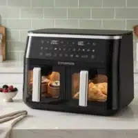 Dyceros Une friteuse noire Dyceros ClearFry Duo 9L (2x4,5L) est posée sur le comptoir d'une cuisine, des muffins cuisant dans le panier de gauche et des pâtisseries dans celui de droite. Son écran numérique est allumé, indiquant les réglages ; un bol de baies repose à proximité.