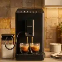 Dyceros Une machine à café noire moderne prépare deux boissons expresso dans des tasses en verre, avec un récipient à lait relié par un tube sur la gauche, sur un comptoir de cuisine avec un dosseret en carrelage et un éclairage chaleureux.
