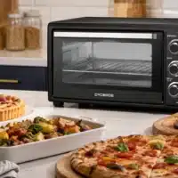 Dyceros Un four de comptoir noir trône sur un plan de travail de cuisine, entouré d'une pizza, d'un plat de légumes, d'un rôti de viande et d'une tarte aux fruits. À l'arrière-plan, on aperçoit des objets de cuisine tels que des bocaux et des plantes.
