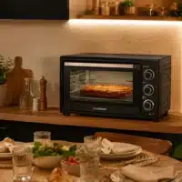 Dyceros Un four de comptoir, dans lequel se trouve un plat au four, est posé sur un plan de travail en bois. Au premier plan, une table à manger est dressée avec des assiettes, des verres, de la salade et du pain. Un éclairage chaleureux crée une ambiance conviviale.