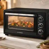 Dyceros Un four grille-pain noir est en train de cuire un plat de légumes, notamment du brocoli et des pommes de terre, posé sur un plan de travail de cuisine, à proximité duquel se trouvent divers ustensiles et des plantes.