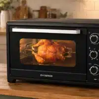 Dyceros Un poulet rôti succulent cuit à l'intérieur du Four électrique Dyceros Solaro 45L, un four de comptoir noir aux lignes épurées, doté de boutons de réglage et d'une vitre à double épaisseur, posé sur un plan de travail en bois. À l'arrière-plan, des plantes, des bocaux et des ustensiles viennent parachever l'ambiance chaleureuse de la cuisine.