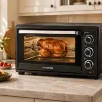 FE450_poulet_roti Dyceros Un poulet entier rôtit à la broche à l'intérieur du Four électrique Dyceros Solaro 35L, équipé d'un système de cuisson par convection et d'une porte en verre à double épaisseur. Cet élégant four de comptoir noir est posé sur un plan de travail de cuisine, à proximité d'une assiette de nourriture et de tomates, tandis que l'on aperçoit des placards de cuisine en arrière-plan.